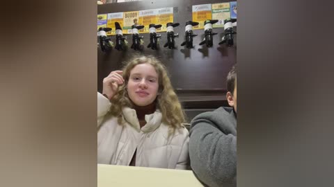 Media: Video of a young girl with curly blonde hair, wearing a white puffy coat, sitting in a restaurant with a dark wooden booth. She's smiling and holding her hair, while behind her, a row of black microphones is visible on a wooden wall.