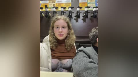 Media: Video of a young woman with curly blonde hair, wearing a brown turtleneck sweater and white jacket, seated in a dimly lit studio with microphones and a poster in the background.