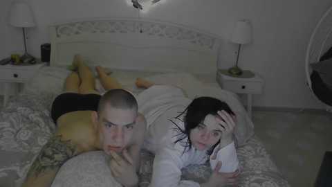 A video depicts a bald man in black shorts lying on a bed with a woman in a white robe, both looking at a smartphone. The room is dimly lit, with white bedding and furniture.