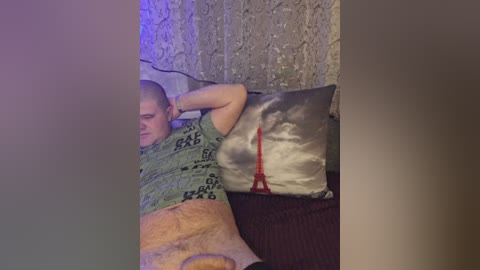 Video of a bald man with fair skin, wearing a green t-shirt and beige shorts, lying on a dark couch, with a gray and silver pillow and a red garment visible.