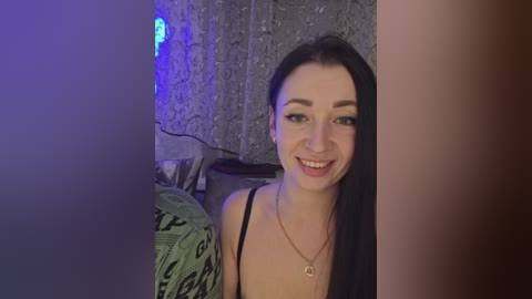 A video of a smiling, young woman with straight black hair, light skin, and a black necklace, standing in a dimly-lit bedroom with gray curtains and a green patterned blanket.