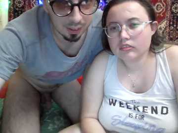 A video of a light-skinned woman with glasses and a white tank top reading \"Weekend Is For...\" kneeling next to a light-skinned man with glasses and a gray shirt, both indoors on a patterned carpet.