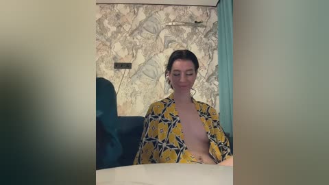 Media: A video of a nude woman with light skin and brown hair, wearing a yellow-patterned kimono, seated in a teal chair, against a textured wall with a decorative mural.