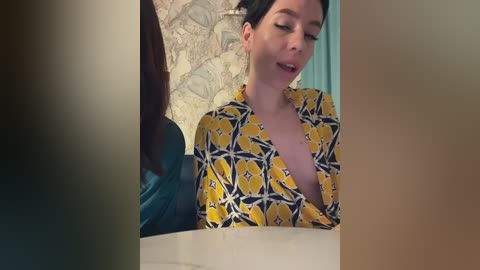 Media: Video of a woman with short black hair, wearing a yellow and black patterned robe, looking surprised in a bathroom with a floral wallpaper background.