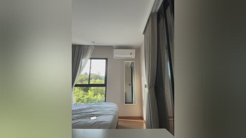 Media: Video of a modern, minimalist bedroom with large windows, light gray walls, and beige curtains. Bed with white sheets and an air conditioning unit on the wall.
