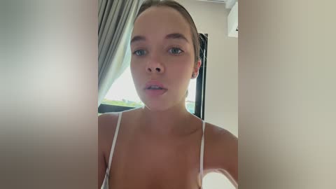 Media: Video of a fair-skinned, young woman with light brown hair in a bun, wearing a white spaghetti-strap top, standing in a brightly lit room with a window and grey curtains in the background.