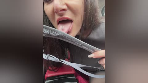 Media: A video of a woman with long, straight brown hair, wearing a gray top and red handbag, sticking her tongue out and holding a ripped car seatbelt.