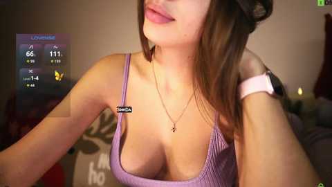 Media: Video of a light-skinned woman with straight brown hair, wearing a low-cut, purple ribbed tank top revealing ample cleavage. She's in a dimly-lit bedroom, with a smartphone displaying a \"LIVE\" status on the left.