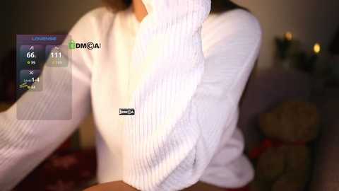 Media: Video of a woman wearing a white, ribbed knit sweater with a black logo, her face obscured. The background is dimly lit with indistinct shapes, possibly furniture.