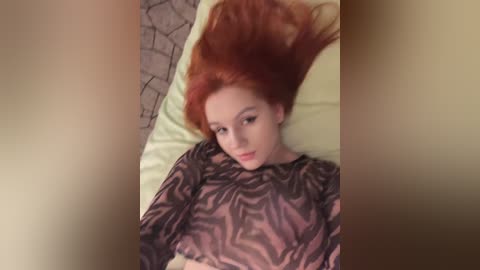 Media: Video of a pale-skinned, red-haired woman with long hair spread on a pillow, wearing a sheer black and white patterned top, lying on a bed with a beige and brown geometric-patterned blanket.