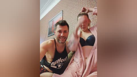 Media: Video of a man and woman, both smiling, in a bedroom with patterned wallpaper and framed picture. The woman is in a black bra and pink robe, while the man wears a black tank top.