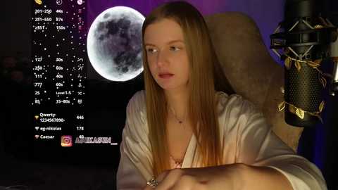 Video of a pale-skinned, long-haired woman in a satin robe, sitting in front of a moon-themed backdrop, with a microphone.