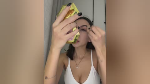 Media: A video of a young woman with fair skin and shoulder-length black hair, wearing a white tank top, taking a bite out of a yellow-wrapped hamburger while holding it in front of her face, partially obscuring her view.
