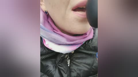Video of a close-up of a person with light skin wearing a colorful, tie-dye scarf, a black puffy jacket, and a microphone, suggesting a recording or podcast setup.