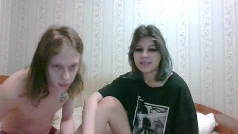 Media: Video of two young women with long, straight brown hair, one topless and one wearing a black t-shirt with a graphic design, sitting on a bed with white floral wallpaper in the background.