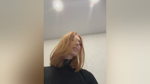 Media: Video of a fair-skinned, red-haired woman in a black turtleneck, smiling slightly, with her head tilted back. The background is a plain, white-walled room with a ceiling light.