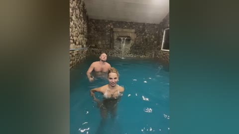 Media: Video of a nude woman and man swimming in a stone-walled, turquoise pool with cascading water feature. Both are smiling, water splashing around them.