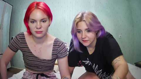 Media: Video of two women with vibrant red and purple hair, sitting on a bed in a room with light green walls and white bedding.