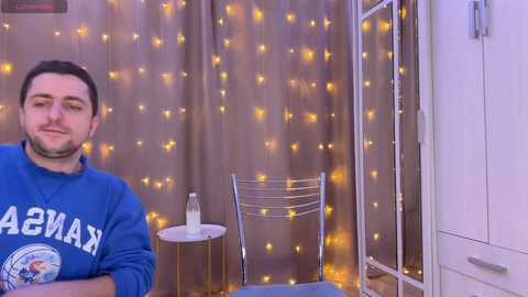 Media: Video of a young man with short dark hair, wearing a blue sweatshirt, sitting at a wooden table with a glass of water, surrounded by a cozy, string-lit curtain backdrop.