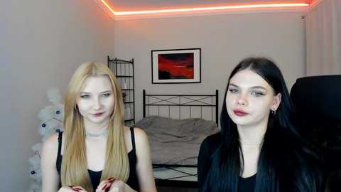 Video of two young women with fair skin, long blonde and black hair, wearing black tops, in a dimly lit bedroom with a bed and red accent lighting.