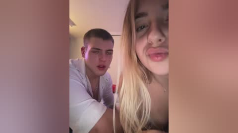 Media: Video of a young Caucasian man with short brown hair, wearing a white robe, and a young Caucasian woman with long blonde hair, puckering her lips, both indoors.