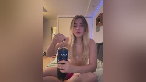 Media: Video of a blonde woman in lingerie, sitting on a bed, holding a Solola soda can, in a dimly lit room with a wooden floor and a closet in the background.