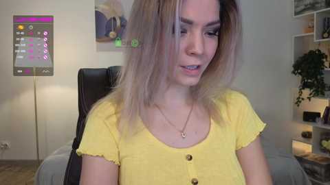 Media: Video of a young woman with light skin and blonde hair, wearing a yellow top, sitting on a black chair in a modern, minimalist bedroom.