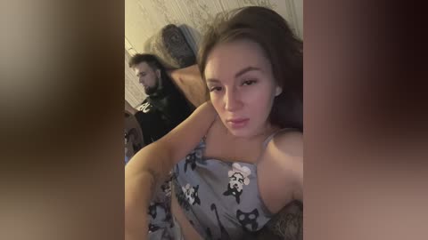 Media: Video of a young, light-skinned woman with brown hair and light makeup, wearing panda-pattern pajamas, taking a selfie. A bearded man with a muscular build and dark hair is partially visible in the background, seated on a chair.