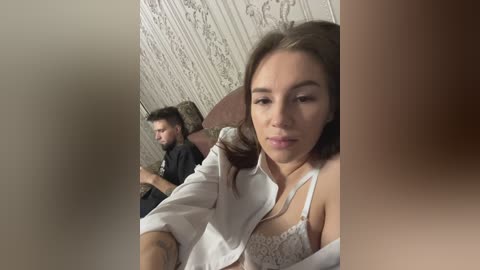 Media: Video of a young woman in a white lace bra, taking a selfie, with a man in the background, both sitting in a room with patterned wallpaper.