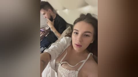 Media: A video showing a young woman with long brown hair, wearing a white lace bralette, taking a selfie in a dimly lit room. A man with short dark hair and tattoos on his arms, dressed in casual clothes, sits in the background.