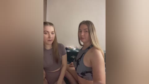 Video of two young women with fair skin and straight hair, wearing casual clothes, standing in a dimly lit room with beige walls and a cluttered dresser in the background.