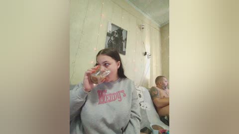 A video of a young woman with long dark hair drinking water in a dimly lit room, wearing a grey Michigan State hoodie. Behind her, a baby sleeps in a crib, adorned with a white blanket and a stuffed toy.