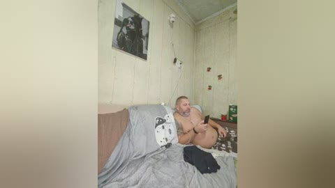 Video of a nude, overweight, middle-aged man with a beard, lying on a bed with gray sheets in a small, cluttered room with white wallpaper, a framed photo, and a green plant.