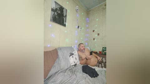 A candid video of a naked man with a muscular build, light skin, and short hair, sitting on a bed with gray sheets, holding a smartphone, surrounded by fairy lights and a black-and-white framed photo on the wall.