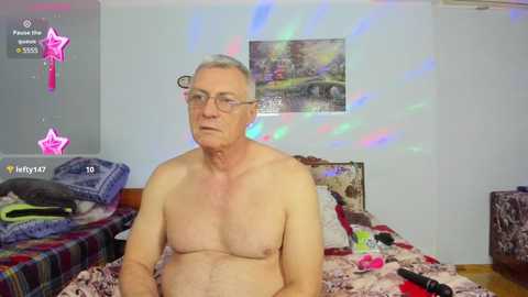 Media: Video of a middle-aged, shirtless, white man with glasses, sitting on a bed with messy clothes and a colorful, abstract light display on the wall.