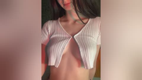 Video of a light-skinned woman with long, dark hair, wearing a cropped, ribbed white top that exposes her small breasts. The background is blurred, featuring soft lighting.