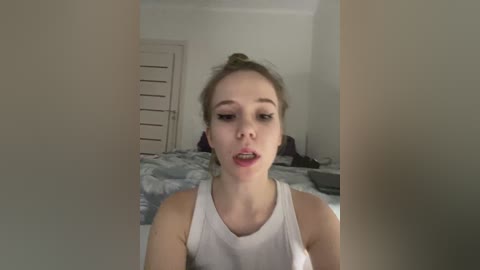 Video of a young woman with fair skin and light brown hair tied in a bun, wearing a white tank top, standing in a bedroom with a white bedspread and closed white door.