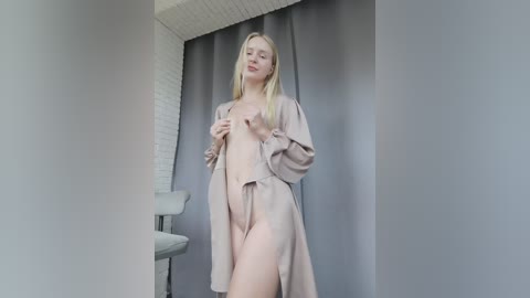 Video of a blonde, slim, Caucasian woman with fair skin, wearing a light beige robe, standing against a grey curtain in a minimalistic room. She has her eyes closed and hands near her chest.