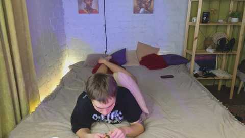 Media: Video of a young man with short brown hair, wearing a black T-shirt, lying on a bed with beige sheets, a red pillow, and a stuffed animal, in a dimly lit, eclectic room with white brick walls and wooden shelves.