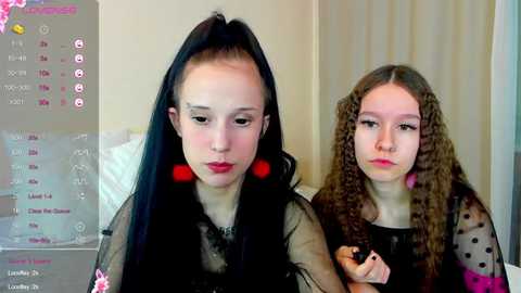 Media: Video of two young girls with fair skin and long, straight hair. One wears a black mesh top and red earrings, the other a black polka-dot top. They stand side by side, looking serious, in a plain, beige room.