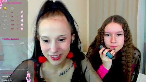 Media: Video of a young woman with a high ponytail and red earrings, smiling, and a young girl with curly hair, holding a phone, in a bedroom.