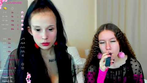 Media: Video of two young girls, one with pale skin, long black hair, wearing a sheer black top, looking sad; another with curly brown hair, wearing a pink sweater, holding a phone, smiling.