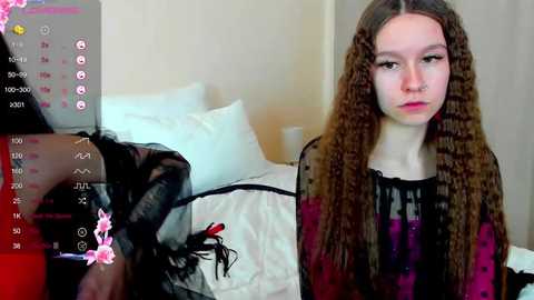 Media: Video of a young woman with long, curly brown hair and pale skin, wearing a black and red patterned dress, sitting on a bed in a dimly lit room.