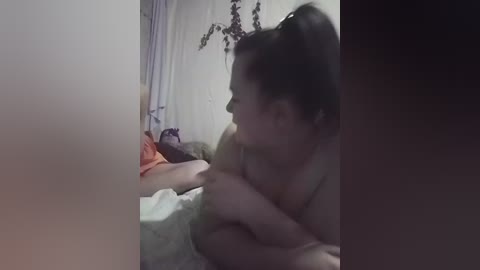 A dimly lit video shows a woman with fair skin, dark hair in a ponytail, and no visible clothing, lying on a bed with white sheets and orange bedding.