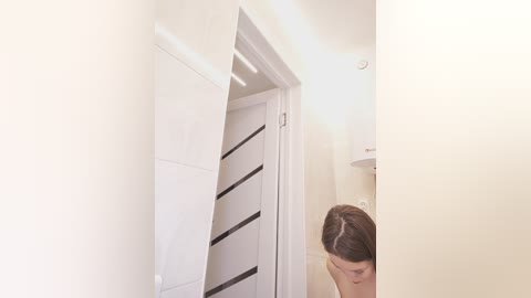 Media: Video of a narrow, well-lit hallway with white walls, a partially open white door revealing a black-striped interior, and a person with shoulder-length brown hair in the background.