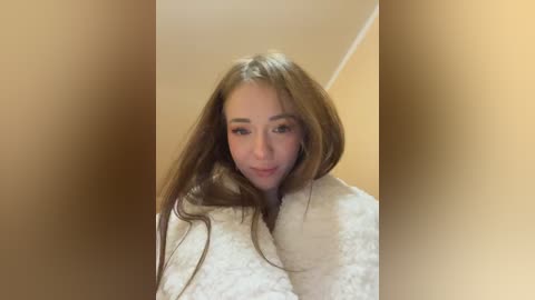 Media: A video of a young woman with long, straight brown hair, wearing a fluffy, white bathrobe, standing indoors against a beige background.