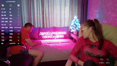 Video of a woman with long brown hair, wearing glasses and a red sweater, seated on a couch, watching a man in an orange shirt and shorts on a gaming console.