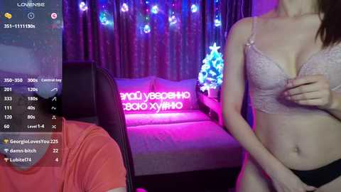 A video of a slender woman in a lacy bra, standing in a room with a Christmas tree and a sofa, with a video game screen overlay.