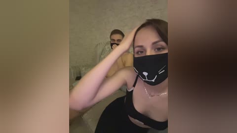 Video of a woman with fair skin, dark hair, wearing a black face mask and a black bra, taking a selfie while a man with short brown hair, wearing a black mask, stands behind her in a dimly lit room.