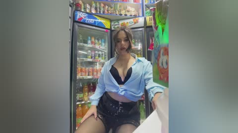 Media: A video of a young woman in a grocery store fridge section, wearing a blue shirt tied at the waist, black bra, and high-waisted shorts. The fridge is stocked with beverages and snacks.
